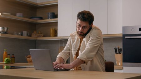 Kaukasischer Multitasking-Mann mit Laptop, der von zu Hause aus telefoniert, freiberuflicher Geschäftsmann in Quarantäne. Männlicher Kunde bestellt Essenslieferung mit Computer-App und Smartphone in der Küche