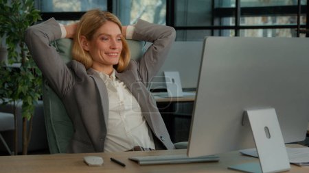 Téléchargez les photos : Souriant calme détendu 40s femme d'affaires fini travail informatique étirement assis au bureau dans le bureau moderne. Heureuse femme d'affaires d'âge moyen se sentant satisfaite après un travail en ligne mettant les mains derrière la tête - en image libre de droit