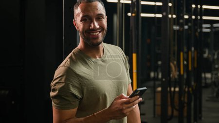 Téléchargez les photos : Africain Américain fitness homme athlète sport entraîneur sourire rire lecture drôle nouvelles regarder humour vidéo blague dans smartphone sportif navigation téléphone mobile rire en regardant la caméra sourire dans la salle de gym - en image libre de droit