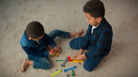 Photo for Top view two little siblings boys African American friends kids schoolboys children play game sit on carpet in living room at home playing with plastic toys repair tools hummer screwdriver spanner - Royalty Free Image