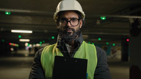 Photo for Portrait Indian man worker contractor mechanic guy in helmet and glasses posing with clipboard folder indoors parking factory warehouse manufacture. Arabian male builder engineer fitter in hard hat - Royalty Free Image