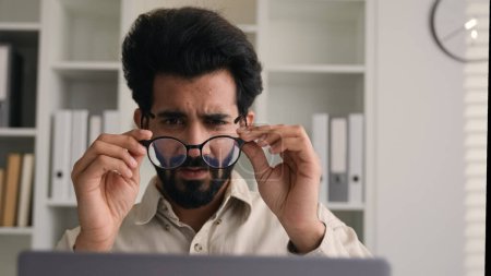 Tired overworked man Indian Arabian businessman working on laptop in glasses confused business employer taking off eyeglasses eyes problem blurry vision check eyesight eye laser correction health care