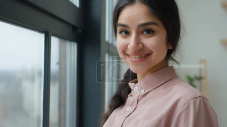 Satisfied girl dreamy Arabian woman looking through window in apartment waiting happy young female businesswoman calm lady dreaming peaceful morning contemplate turns to camera smiling at home office