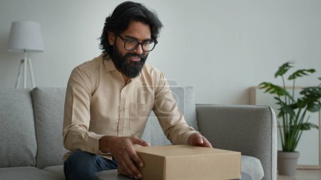Téléchargez les photos : Heureux client masculin client consommateur Arabe homme indien gars dans des lunettes boîte ouverte en carton à la maison déballer achats en ligne commander paquet enveloppé obtenir livraison cadeau déballage carton colis - en image libre de droit
