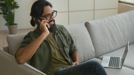 Téléchargez les photos : Musulman arabe gars pigiste assis sur canapé avec ordinateur portable parler téléphone mobile à la maison souriant homme d'affaires indien dans les lunettes parler smartphone répondre appel insouciant converser cellule discussion d'affaires - en image libre de droit