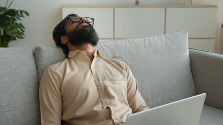 Foto de Cansado freelancer agotado trabajando con el ordenador portátil en casa sobrecargado de trabajo árabe musulmán hombre se duermen en el sofá enfermo cansado durmiendo indio hombre de negocios se apoyan en el sofá cansancio fatiga necesidad descanso relajarse - Imagen libre de derechos