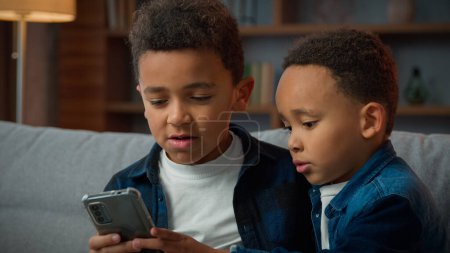 Photo for Two siblings African American brothers sons children boys friends little kids at home using mobile phone without parental control browsing smartphone concentrate with online video game in cellphone - Royalty Free Image