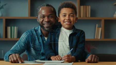 African American happy family portrait smiling at camera little boy child schoolboy with father at table help with homework. Dad smile help son with lesson class kid homeschooling elementary education