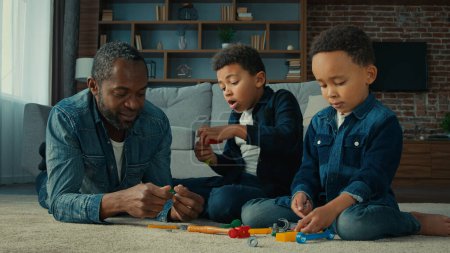 Téléchargez les photos : African American ethnic man father with two little preschool sons boys children play game lying on floor at home dad with kids playing with tools toys talking having fun together enjoy funny weekend - en image libre de droit