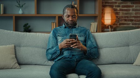 Téléchargez les photos : Seul homme afro-américain gars assis à hone canapé canapé en utilisant le téléphone mobile smartphone chat navigation téléphone portable dans l'appartement achat en ligne app internet chat utilisation des médias sociaux - en image libre de droit