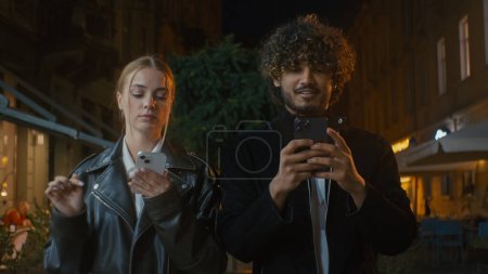 Photo for Couple walking city outside street night European woman and Indian Latino Arabian man texting using smartphone mobile phone multiracial disorder problem frustrated offended girl ignore guy separation - Royalty Free Image