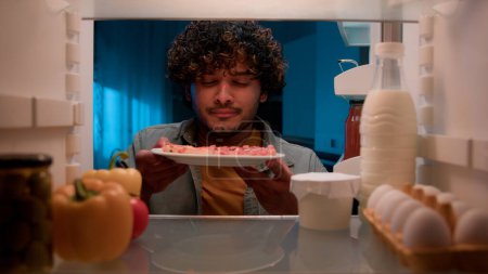 View inside from refrigerator Indian sly man with curly hair open fridge at night evening in kitchen hungry guy male looking around take out slice of pizza overeating at nighttime unhealthy nutrition