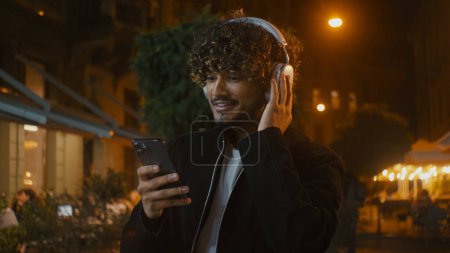 Téléchargez les photos : Indien Latino Arabe heureux homme marche rue de la ville en plein air soir écouter de la musique écouteurs en utilisant smartphone téléphone mobile gadget beau gars élégant sourire masculin satisfait week-end moderne - en image libre de droit