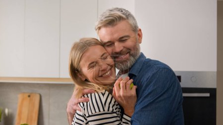 Photo for Adults happy family 40s smiling positive couple middle-aged man woman at kitchen cuddling hug embracing food delivery order wife give apple husband embrace hugging at kitchen cuddle love bonding. High - Royalty Free Image