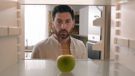 Photo for POV point of view from inside refrigerator Caucasian adult man at kitchen open empty fridge with one green apple fruit hungry guy male upset need food delivery meal crisis diet shrugs sad frustrated - Royalty Free Image