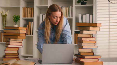 Multitasking busy standing Caucasian pupil high school student girl woman Caucasian lady teenager preparing exam studying learning at home university library with many books writing notes using laptop