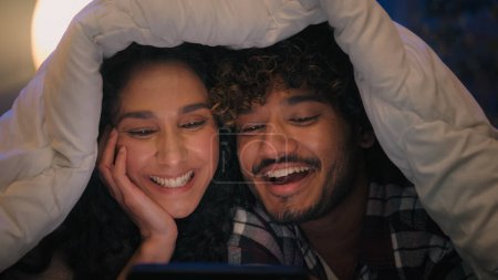 Téléchargez les photos : Multiracial heureux couple insouciant Indien homme Hispanique femme regarder film drôle vidéo rire souriant avoir amusant humour ridicule nouvelles blague téléphone portable rire couverture sous couverture la nuit - en image libre de droit