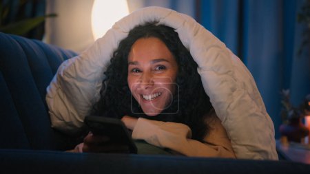 Téléchargez les photos : Happy souriant hispanique insouciante fille latino femme indienne dame Arabe sourire féminin à la caméra la nuit soir sombre maison sous couverture avec couette en utilisant le téléphone mobile influenceur gadget addict médias - en image libre de droit