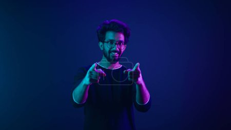 Foto de Retrato en neón ultravioleta feliz indio hombre árabe trabajador codificador hacker ingeniero informático hacer gesto hey que muestra el dedo índice dirección delantera en la cámara pulgares hacia arriba de acuerdo apoyo recomiendo - Imagen libre de derechos