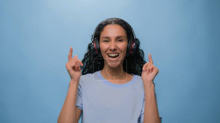 Photo for Studio portrait happy smiling singing Arabian woman Indian lady Hispanic girl listening to music with headphones dancing listen radio dance move at blue background headset sound audio fun song rhythm - Royalty Free Image