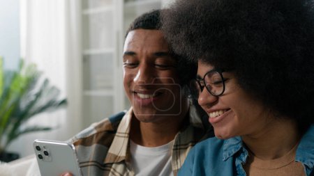 Téléchargez les photos : Couple afro-américain homme et femme ensemble à la maison en regardant le téléphone à l'aide d'un appareil mobile gadget pour rechercher l'offre de choisir des marchandises réservation en ligne billets pour les vacances discuter parler budget familial - en image libre de droit