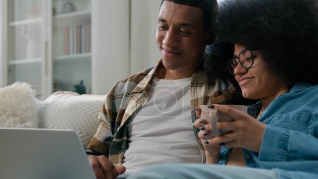 Photo for African American couple at home couch looking at laptop talking watching film movie discuss choosing goods online store internet shopping order delivery using computer together woman drinking tea cup - Royalty Free Image
