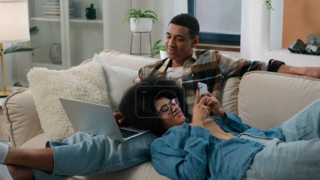 Photo for African American couple man with laptop computer sitting on couch home woman chatting smartphone browsing mobile phone laying on male knees girl guy gadget addict internet addiction smiling family - Royalty Free Image