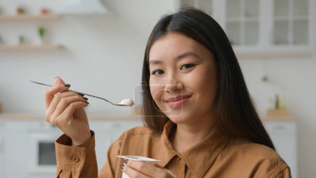 Foto de Asiático coreano mujer chica degustación saludable nutrición comer cremoso fruta yogur en cocina chino chica sonriendo a cámara comer lácteos lactosa postre con cuchara desayuno comida pérdida peso dieta salud - Imagen libre de derechos