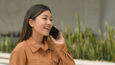 Téléchargez les photos : Asiatique femme heureux client marcher dans le centre commercial parler téléphone réponse appel mobile parler insouciant chinois japonais coréen femme d'affaires va dans entreprise communication cellulaire entreprise avec smartphone - en image libre de droit