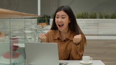Téléchargez les photos : Heureuse choquée femme d'affaires asiatique assis dans un café avec ordinateur portable surprise coréenne fille gagnante gagner offre d'emploi célébrer enchère internet gagner prix en ligne crier se réjouir victoire succès surprise - en image libre de droit