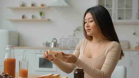 Téléchargez les photos : Asiatique femme au foyer chinois coréen japonais fille mettre sur pain pâte de chocolat appliquer confiture sucrée sur pain grillé femme multiethnique cuisinier petit déjeuner à la maison cuisson sandwich dans la cuisine nourriture de la livraison de supermarché - en image libre de droit