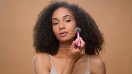 Photo for African American woman applying powder make-up natural beauty clean soft moisturized hydrated skin care treatment girl make blush on face bronze contouring using makeup brush cosmetic beige background - Royalty Free Image