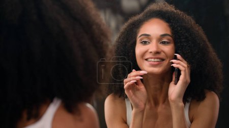 Photo for Beautiful African American woman happy smiling girl looking at mirror reflection touch face make up preparing skin care moisturize with cream skincare procedure talking mobile phone call smartphone - Royalty Free Image