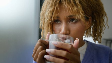 Téléchargez les photos : Ethnique afro-américain heureux souriant inspiré femelle à la maison boire chaud à base de plantes goût sain thé noir regarder la fenêtre femme fille tenant tasse avec boisson se détendre dans l'appartement profiter de rêver matin - en image libre de droit