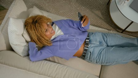 Téléchargez les photos : Afro-américaine fille ethnique couché sur le canapé se détendre sur canapé confortable doux repos relaxant en utilisant l'application de téléphone mobile vérifier les médias sociaux fil chat smartphone navigation téléphone portable maison week-end - en image libre de droit