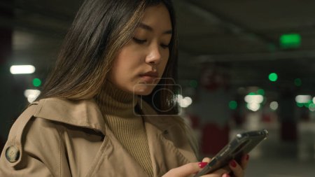 Téléchargez les photos : Sérieux asiatique femme chinoise coréen japonais femme fille ethnique dame client conducteur dans le parking souterrain bavarder application de téléphone mobile dactylographier message navigation internet téléphone portable chat - en image libre de droit