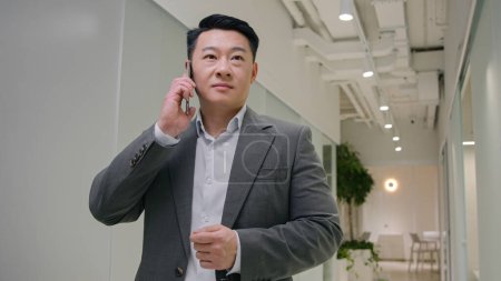 Téléchargez les photos : Heureux Asiatique homme d'affaires chinois japonais homme ethnique appelant téléphone mobile dans le couloir de bureau moderne mâle joyeux employeur gestionnaire d'entreprise réponse smartphone appel discuter converser négocier communiquer - en image libre de droit