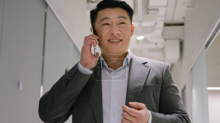 Téléchargez les photos : Heureux Asiatique homme d'affaires chinois japonais homme ethnique appelant téléphone mobile dans le couloir de bureau moderne mâle joyeux employeur gestionnaire d'entreprise réponse smartphone appel discuter converser négocier communiquer - en image libre de droit