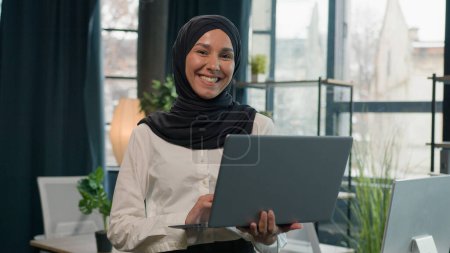 Téléchargez les photos : Islam femme d'affaires en hijab Arabe Indien musulman musulman femme d'affaires entrepreneur gestionnaire fille avec ordinateur portable commerce travailler étudier la navigation en ligne ordinateur sourire dents à la caméra dans le bureau - en image libre de droit