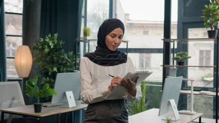 Téléchargez les photos : Heureuse musulmane islam femme Arabe fille dans hijab HR manager secrétaire avec tablette papier écrire des notes signer des données dans la tablette écriture à faire liste des idées d'affaires succès femme d'affaires planification flux de travail - en image libre de droit