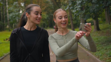 Photo for Two happy Caucasian sport girls bloggers influencers women using mobile phone smartphone call record video blog vlog healthy lifestyle workout in city active smiling female sportswomen walking in park - Royalty Free Image