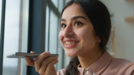 Fröhlich fröhlich arabisch indische Business-Mädchen sprechen Handy in der Nähe des Fensters Geschäftsfrau sagen auf dem Lautsprechertelefon aufnehmen Voice-Mail-Audiobotschaft Anrufbeantworter sprechen am Handy verwenden Tonrekorder