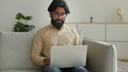 Photo for Arabian muslim man freelancer working from home focused millennial guy indian businessman typing on computer laptop surfing internet looking at screen lying on couch distant remote job on quarantine - Royalty Free Image