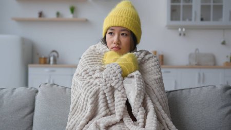 Téléchargez les photos : Congelé asiatique chinois coréen femme malade triste fille congélation assis dans appartement froid souffrir basse température gel gelé recouvert de couverture chaude chapeau et mitaines hiver saison problème de chauffage dans la cuisine à la maison - en image libre de droit