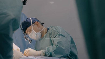 Téléchargez les photos : Chirurgien homme praticien avec des assistants d'équipe effectuer une opération chirurgicale médecin couture sutures de plaie peau avec des outils médicaux clips fil dans la salle d'opération de l'hôpital patient traitement de santé chirurgie - en image libre de droit