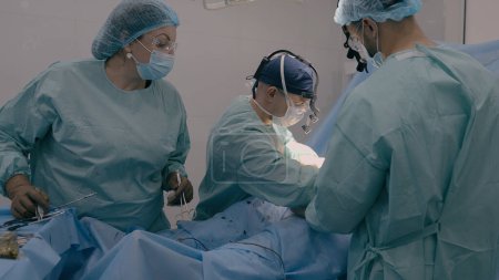 Téléchargez les photos : Chirurgien avec l'homme assistant médecin opératoire patient effectuer une intervention chirurgicale. Infirmière femme debout à table avec des instruments médicaux découpés appendicite tumeur maligne processus de chirurgie cardiopathie - en image libre de droit