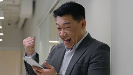 Photo for Excited guy in hallway office business man middle-aged entrepreneur Asian chinese korean multiethnic businessman winning mobile phone bet app online lottery male winner win scream shout achievement - Royalty Free Image