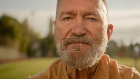 Photo for Close up old Caucasian man pensioner looking at camera serious face expression calm silently blinking athlete recreation stadium outside city relax sportsman bearded wrinkled male wellbeing lifestyle - Royalty Free Image