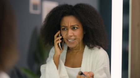 Téléchargez les photos : Colère émotionnel frustré stressé afro-américain femme agacé irritée ethnique fille dame poudre visage bio cosmétique soins de la peau regarder miroir à la maison salle de bain parler conflit téléphonique - en image libre de droit