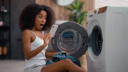 Afro-americana ama de casa triste disgustada biracial chica étnica sacar de la lavadora de lavandería tomar pantalones vaqueros encontrar roto húmedo teléfono inteligente olvidado teléfono móvil en el bolsillo de tela gadget impermeable